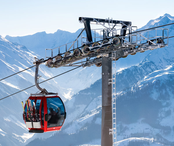 The image shows a red cable car gondola traveling along a steel cable over a snowy mountain landscape.